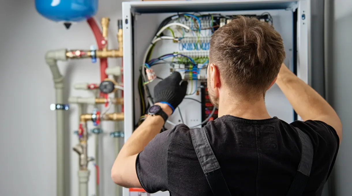 Technician working on the wiring inside an electrical control panel next to building pipework