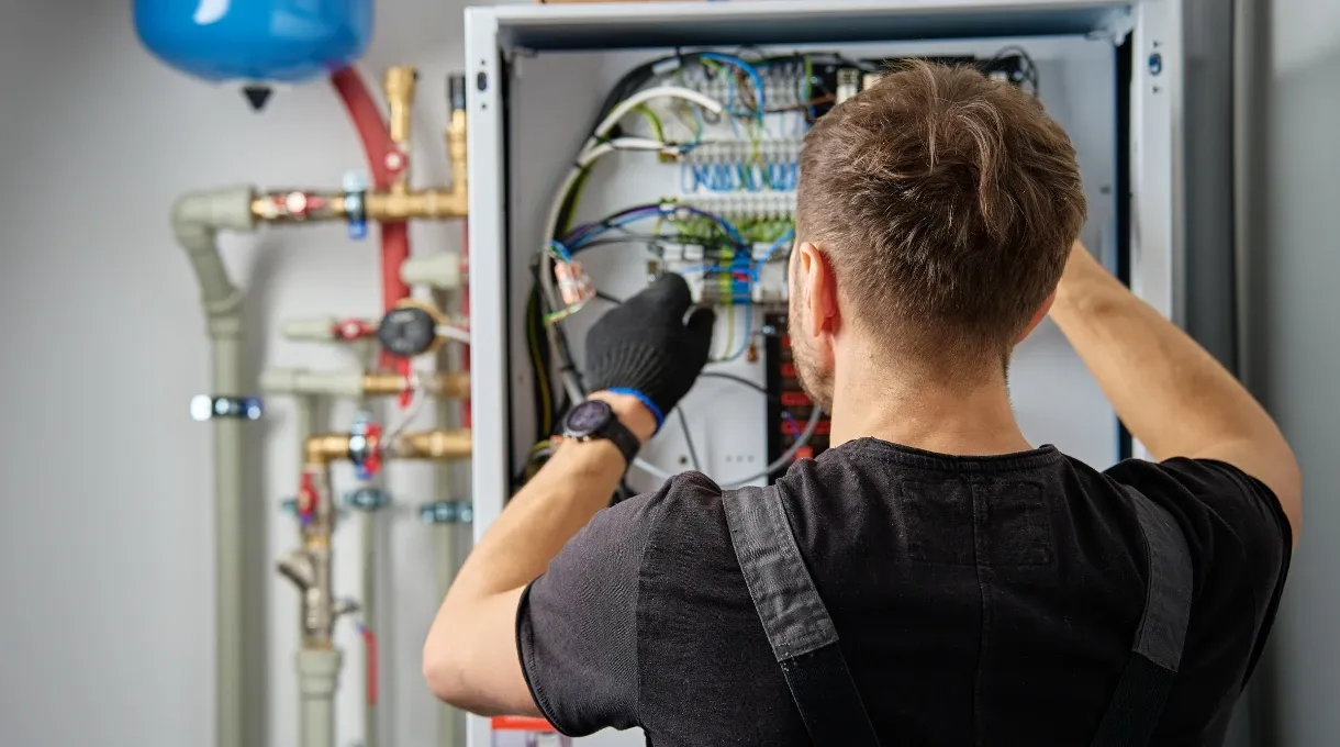 Technician working on the wiring inside an electrical control panel next to building pipework